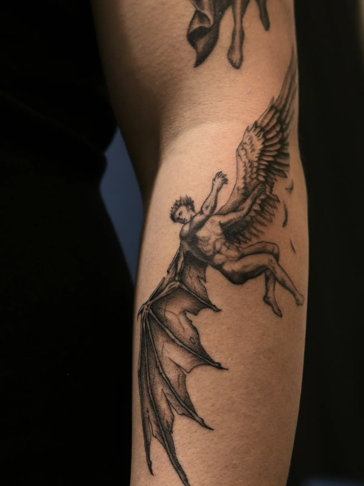 Fall of Lucifer black and grey tattoo on the outer forearm, showing a winged figure transforming from feathered to bat-like wings, Prague by Andrey Kovalov