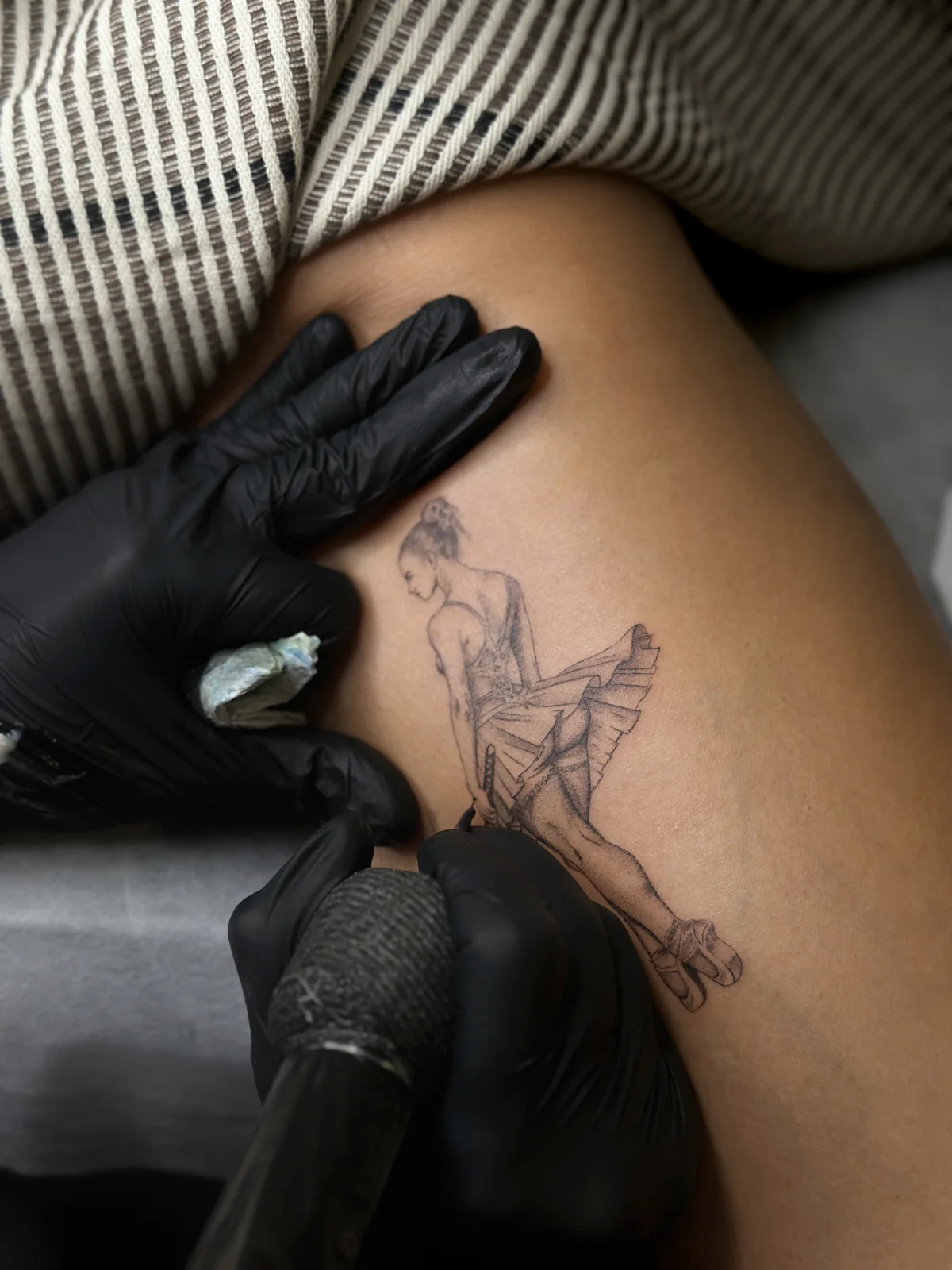 Ballerina with katana thigh tattoo during the tattooing process, with the artist's gloved hands and machine visible, Prague by Andrey Kovalov
